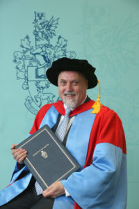 Stefan Cross, seated in graduation regalia, holding his Honorary Degree