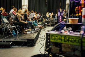 The orchestra rehearsing backstage with an amplifier in the foreground reading DON BROCO