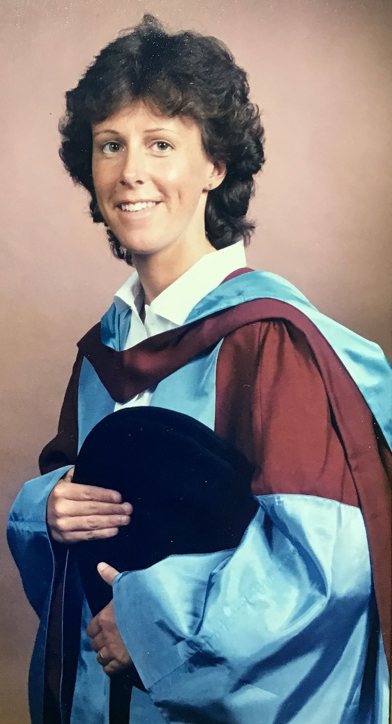 Penny Phillips as a successful PhD graduate, in regalia and smiling to camera, her hat under her arm